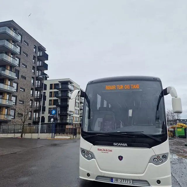 Hvit turbuss fra Risør Tur & Taxi parkert foran moderne leilighetsbygg på overskyet dag.