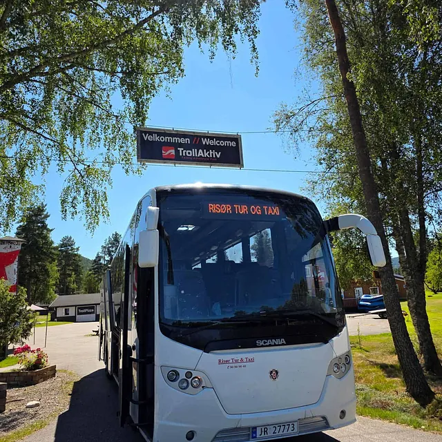 Hvit turbuss fra Risør Tur og Taxi står parkert ved TrollAktiv under solfylte trær.
